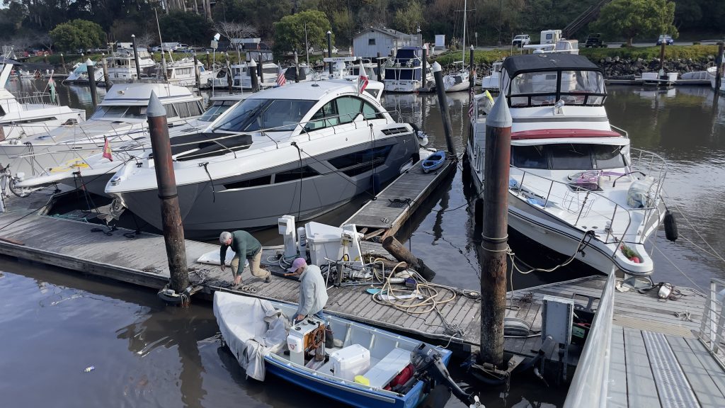 Dock with two large boats tied up on one side and one small boat tied up on the other side. One person in the small boat. One person crouching on the dock next to the small boat