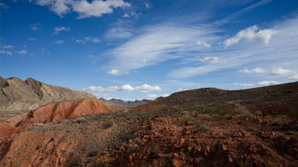 Landscape with blue sky and white clouds, mountains in the distance, red rocky ground in foreground